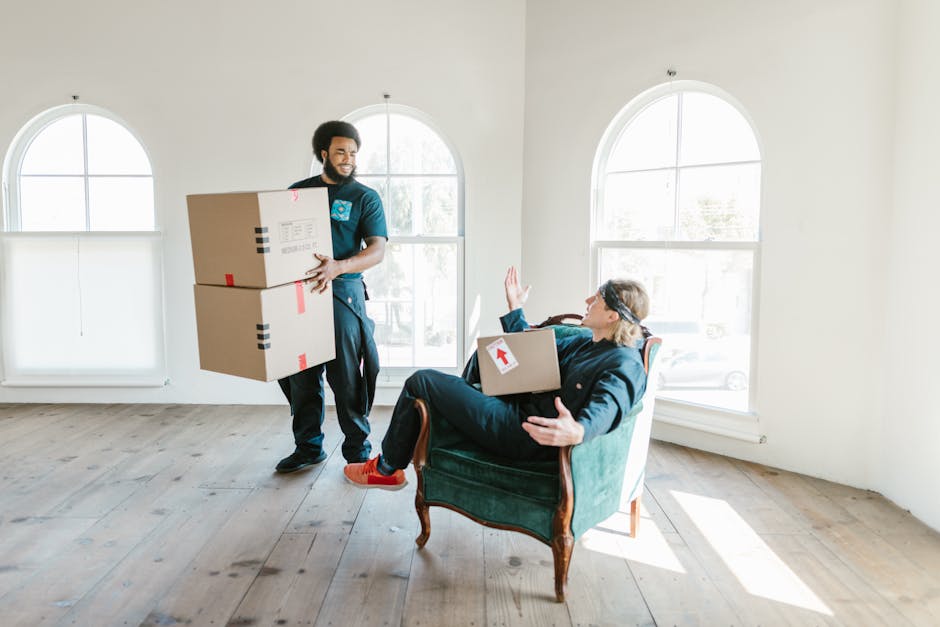 A young woman sitting cross-legged on a wooden floor inside a room, surrounded by multiple large cardboard moving boxes of various sizes, some sealed with red packing tape and labelled with black prin