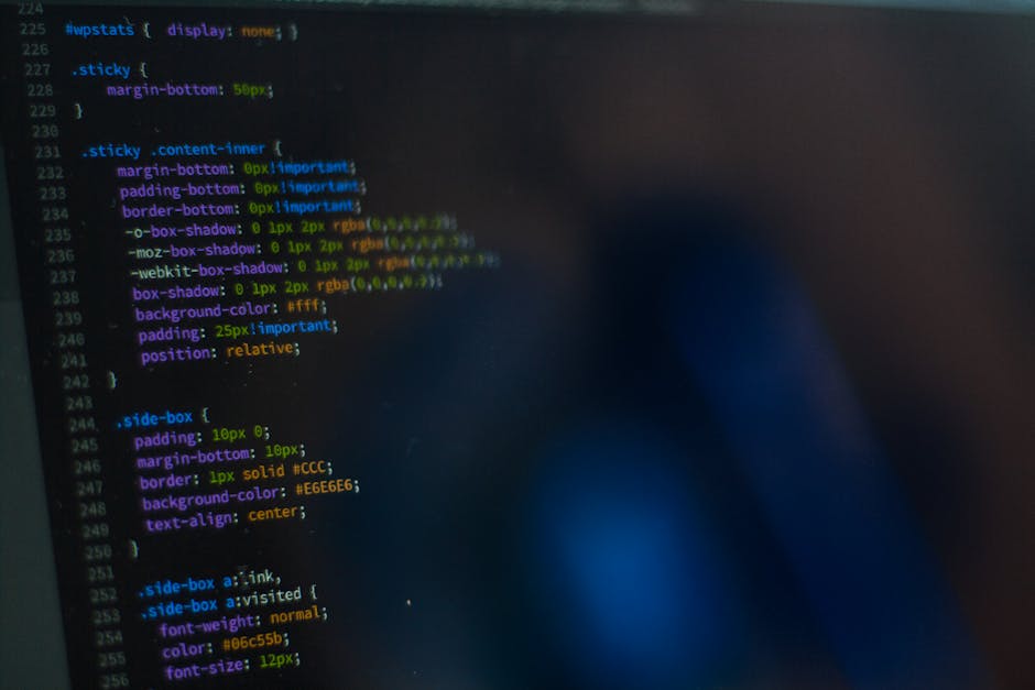 A computer monitor displays a code editor with lines of JavaScript code highlighted in various colours, including pink, blue, and green, on a dark background. The screen is tilted slightly, showing th